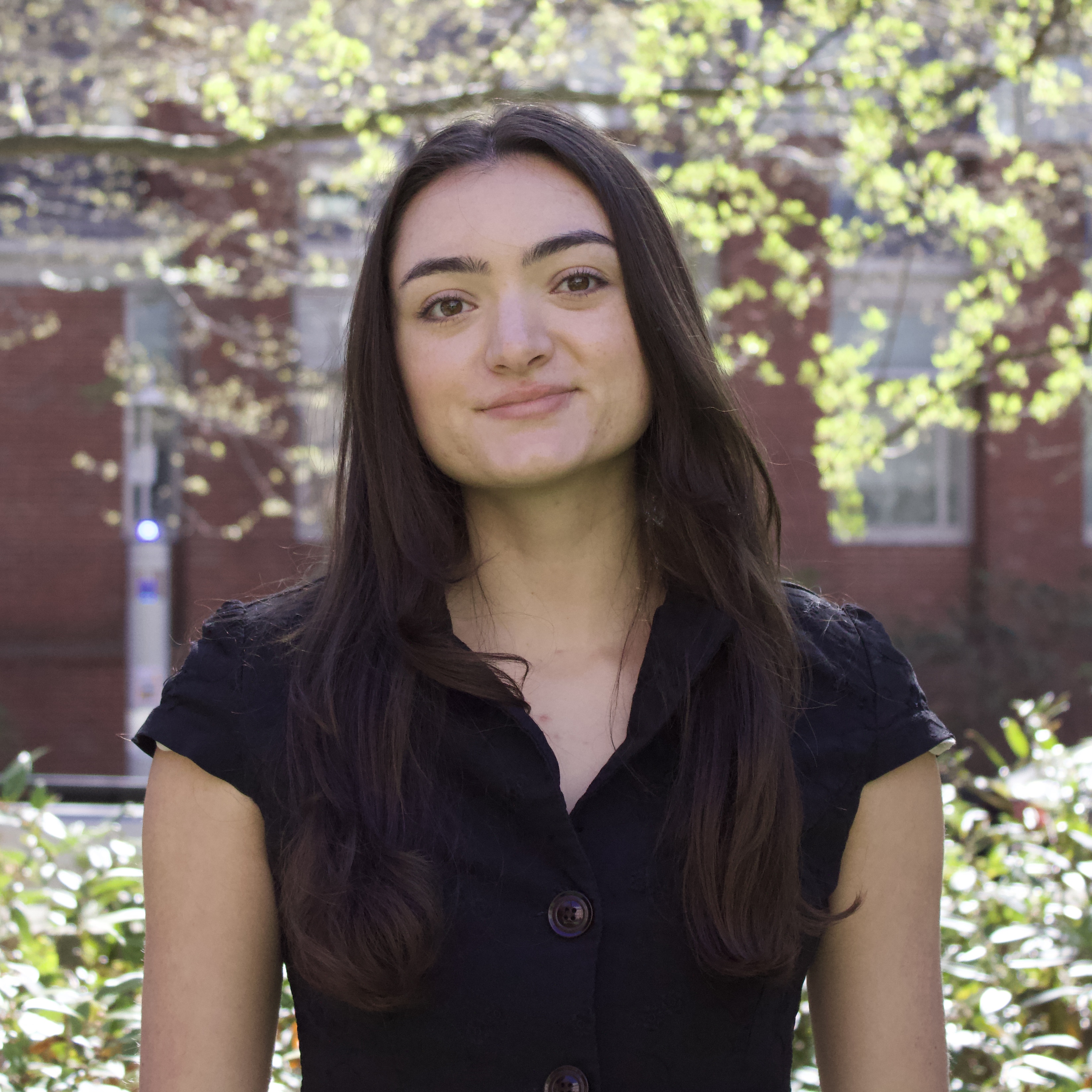 Karina outside Hegeman Hall at Brown University. She has long dark brown hair and is wearing a black collared blouse.